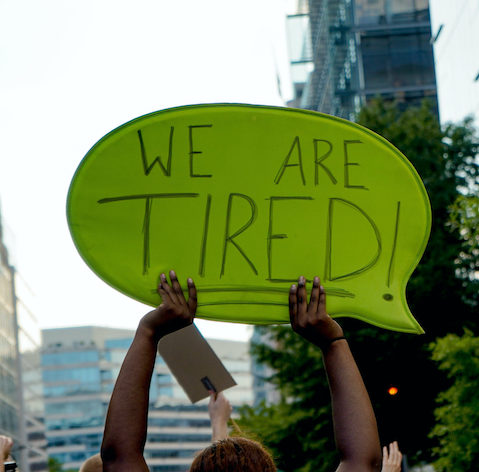 Black woman protesting with a sign that reads We are TIRED!