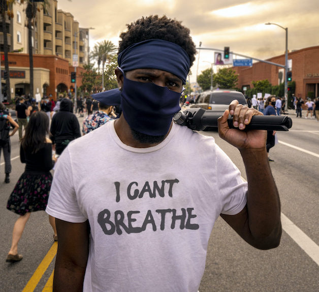 Black man in face mask wearing a shirt that reads I Can't Breathe and holding a camera tripod